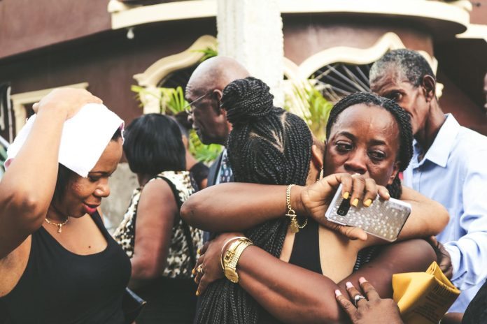 Unhappy African American female with smartphone embracing unrecognizable friend near people on street